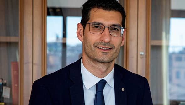 Man wearing glasses and a suit, seated in front of a wooden cabinet with books and decorative items. - Olive Oil Times