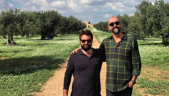 Two men posing together in an olive grove with trees and grass in the background. - Olive Oil Times