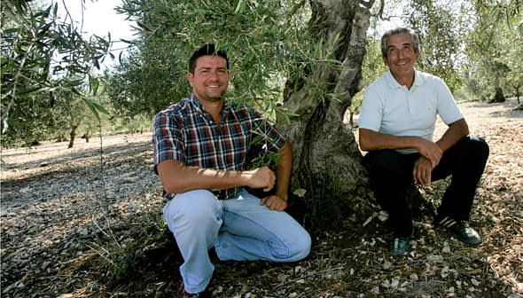 Two men kneeling beside an olive tree in an olive grove, both smiling at the camera. - Olive Oil Times