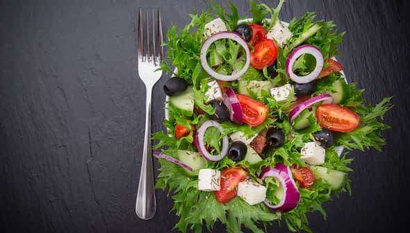 A colorful garden salad featuring mixed greens, tomatoes, cucumbers, olives, and feta cheese on a dark surface. - Olive Oil Times