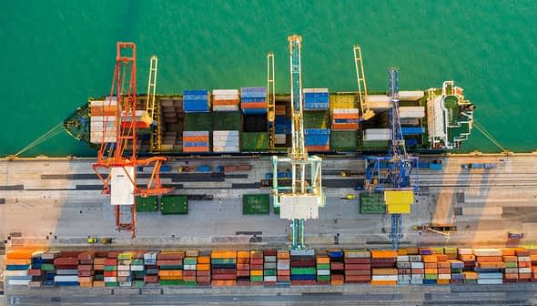 Aerial view of a cargo ship docked at a port with cranes and stacked containers. - Olive Oil Times