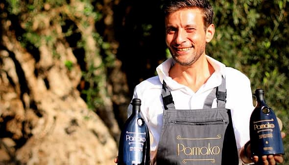 Man in an apron holding two bottles of Pamako olive oil outdoors with trees in the background. - Olive Oil Times