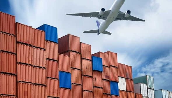 An airplane flying above a stack of colorful shipping containers at a port. - Olive Oil Times
