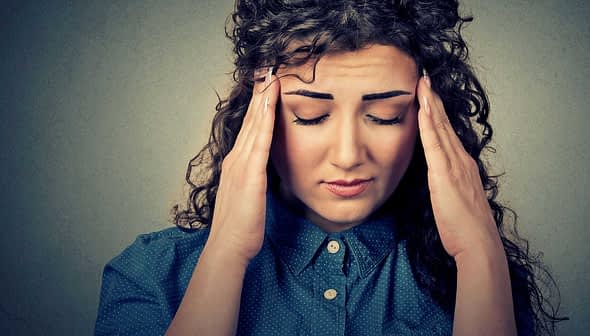 A woman with curly hair wearing a blue shirt, holding her head with both hands and closing her eyes. - Olive Oil Times
