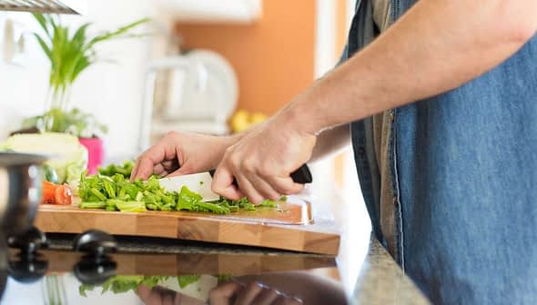 A person using a knife to chop green vegetables on a wooden cutting board in a kitchen. - Olive Oil Times