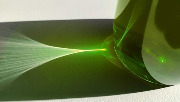 Green glass bottle casting a light reflection on a white surface. - Olive Oil Times