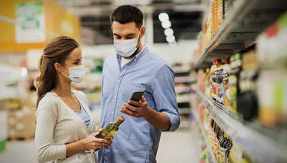 A man and woman wearing masks while shopping in a grocery store aisle, examining products. - Olive Oil Times