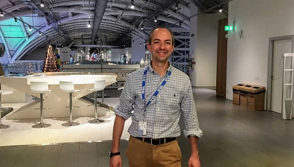 A man wearing a checked shirt and beige pants standing in a modern indoor area with a bar-like setup. - Olive Oil Times