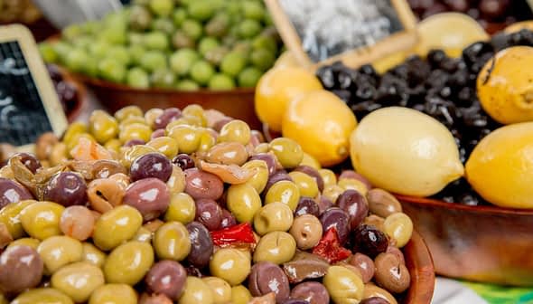A bowl filled with various types of olives alongside lemons at a market display. - Olive Oil Times