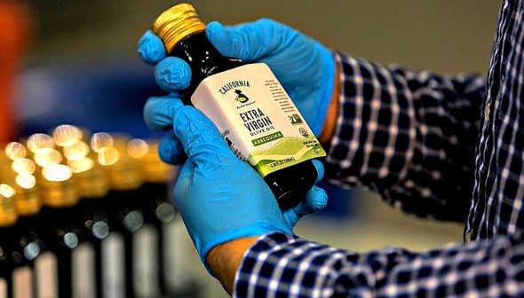 Person wearing blue gloves holding a bottle of California extra virgin olive oil with a label. - Olive Oil Times