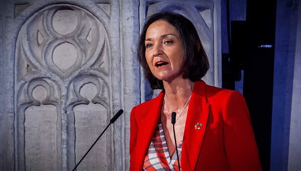 Woman in a red blazer speaking at a podium with a decorative background. - Olive Oil Times