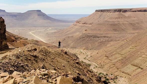 Individual standing on a rocky outcrop overlooking a vast desert landscape with distant mountains. - Olive Oil Times