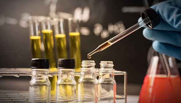 A laboratory scene showing a hand in a blue glove using a pipette to transfer liquid into small glass bottles. - Olive Oil Times