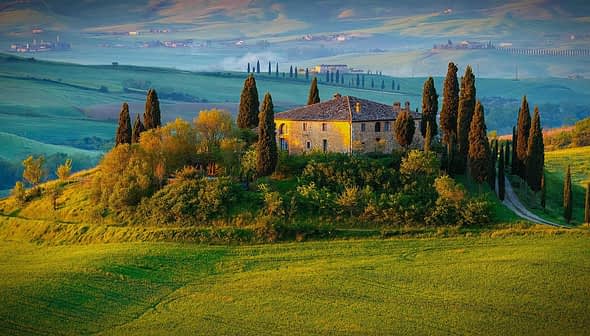 Stone house surrounded by cypress trees on a hill in a Tuscan landscape with rolling green fields. - Olive Oil Times