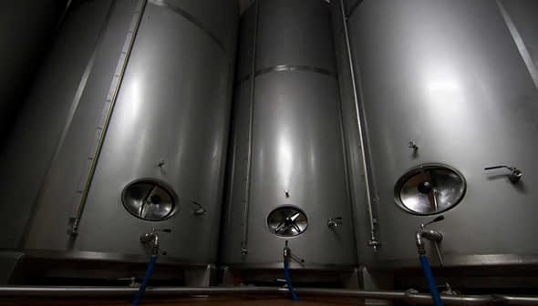 Three stainless steel storage tanks with valves and pipes in a facility. - Olive Oil Times
