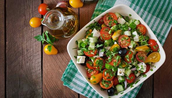 A bowl of fresh vegetable salad with tomatoes, cucumbers, olives, and herbs on a wooden table. - Olive Oil Times
