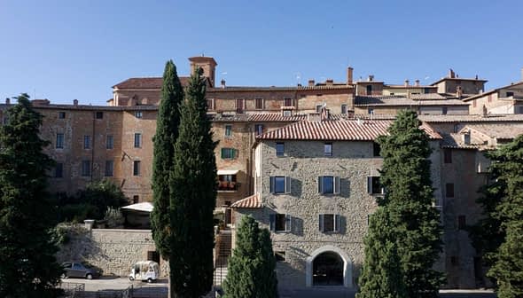 Stone buildings with terracotta roofs in a Tuscan village, surrounded by tall trees. - Olive Oil Times