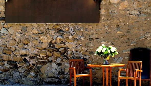 A wooden table with two chairs and a vase of flowers on a stone wall background. - Olive Oil Times