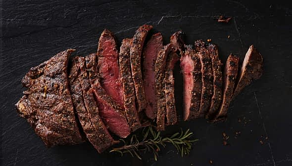 Sliced roast beef arranged on a slate surface with herbs. - Olive Oil Times