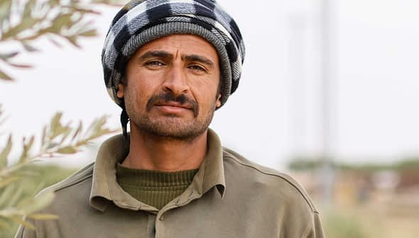 A man wearing a traditional black and white checked headscarf and a green shirt, standing outdoors. - Olive Oil Times