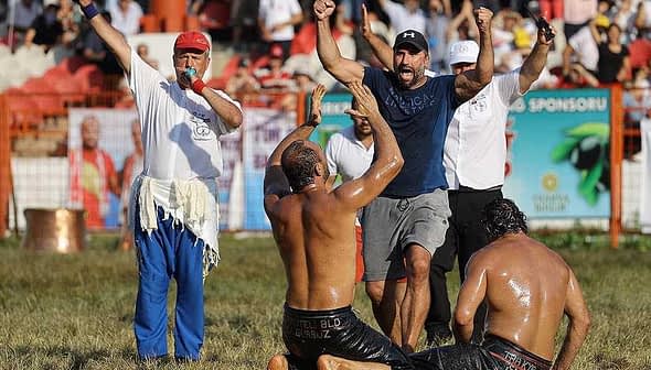 Two men wrestling on the ground while a referee and spectators cheer in the background. - Olive Oil Times