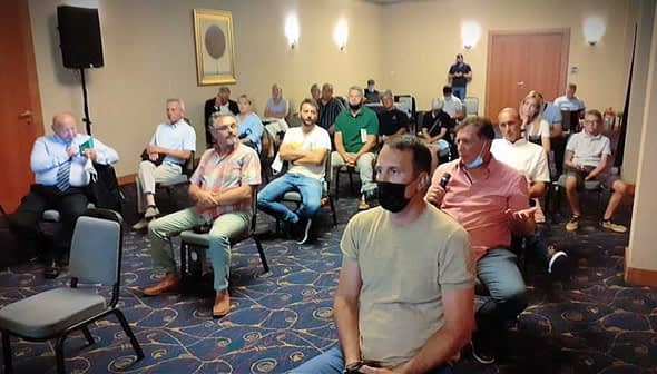 Group of people seated in a meeting room, some wearing masks, listening to a speaker. - Olive Oil Times