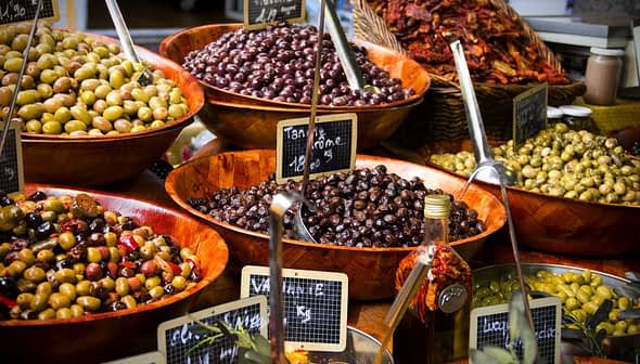 Assorted olives displayed in wooden bowls at a market, with labels indicating prices. - Olive Oil Times