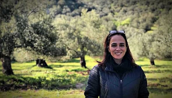 A woman with dark hair and sunglasses standing in an olive grove with trees in the background. - Olive Oil Times