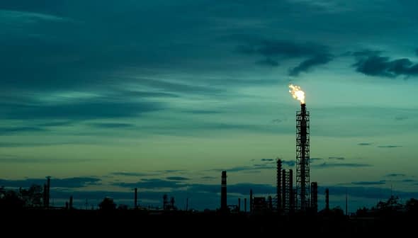 Flare stack emitting flame against a dusk sky with clouds and silhouettes of industrial structures. - Olive Oil Times