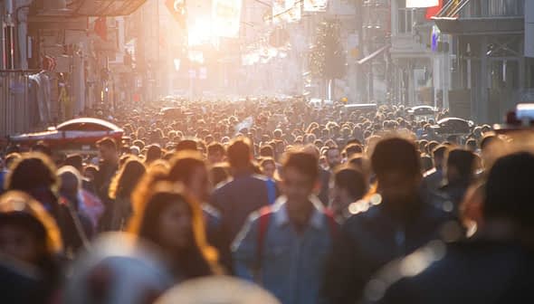 A large crowd of people walking in a busy urban street during sunset with sunlight filtering through. - Olive Oil Times