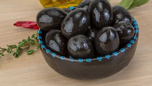 A bowl filled with black olives on a wooden surface with herbs and spices nearby. - Olive Oil Times