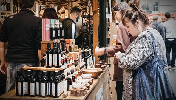 Various bottles of olive oil displayed on a wooden table at a market with people in the background. - Olive Oil Times