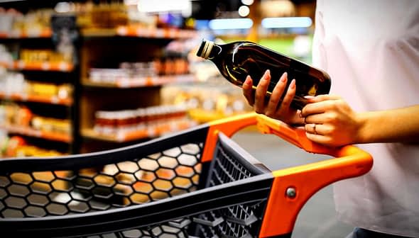 Individual holding a bottle of olive oil while standing next to a shopping cart in a store. - Olive Oil Times