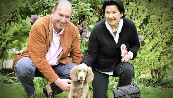 A man and a woman kneeling beside a light brown puppy in a garden setting. - Olive Oil Times