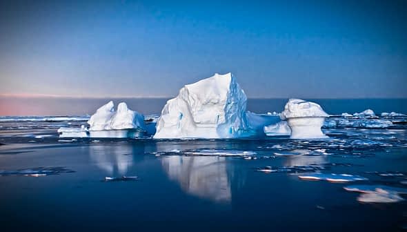 Icebergs floating in calm waters with a blue sky in the background during twilight. - Olive Oil Times