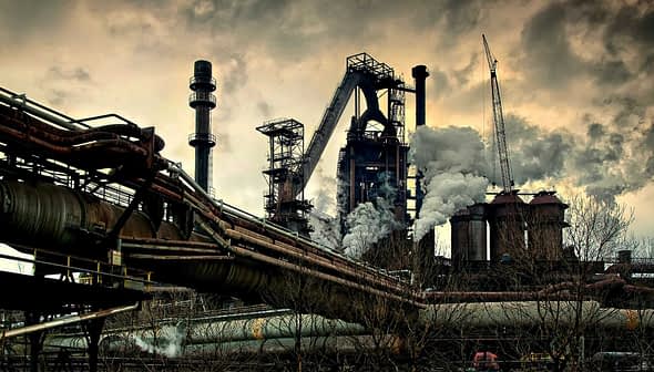Industrial steel mill featuring large smoke stacks and pipes with visible emissions. - Olive Oil Times