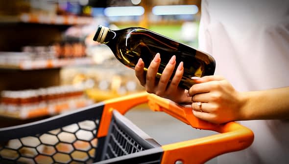 Person holding a bottle of olive oil while pushing a shopping cart in a store. - Olive Oil Times
