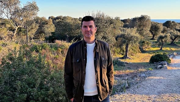 A man wearing a dark jacket and white sweater standing in an olive grove with trees in the background. - Olive Oil Times