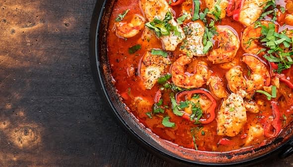A black dish containing chicken pieces, red peppers, and herbs in a tomato-based sauce. - Olive Oil Times