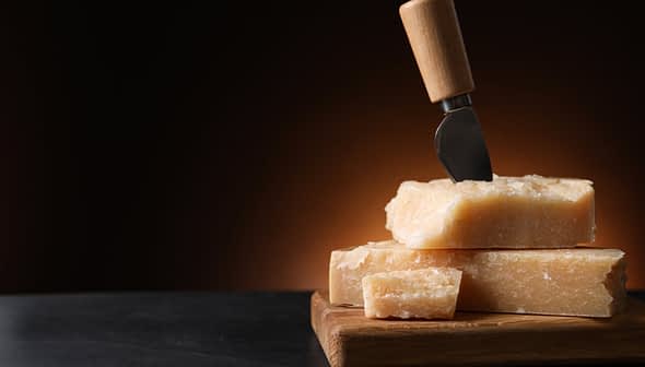 Block of Parmesan cheese with a cheese knife inserted into it, resting on a wooden cutting board. - Olive Oil Times