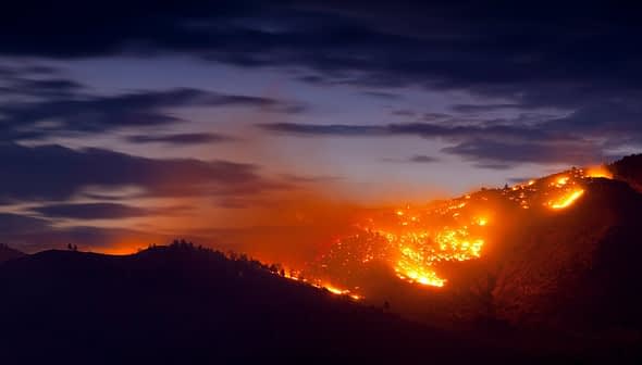 A mountain landscape with a wildfire burning along the slope under a twilight sky. - Olive Oil Times