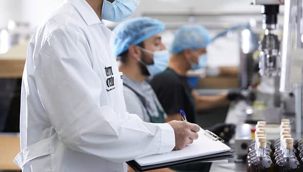Three workers in a food processing facility wearing masks and hairnets, with one taking notes on a clipboard. - Olive Oil Times