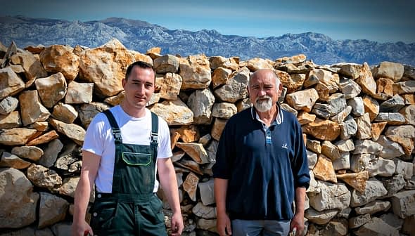 Two men posing in front of a stone wall with mountains in the background. - Olive Oil Times