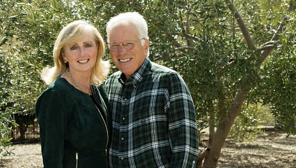 A couple smiling while posing outdoors in front of olive trees, wearing casual clothing. - Olive Oil Times