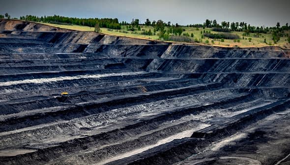 Aerial view of an open-pit mining site with layered black earth and machinery in the distance. - Olive Oil Times