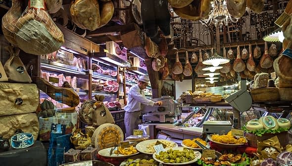 Interior of an Italian deli featuring various meats, cheeses, and a staff member slicing meat. - Olive Oil Times