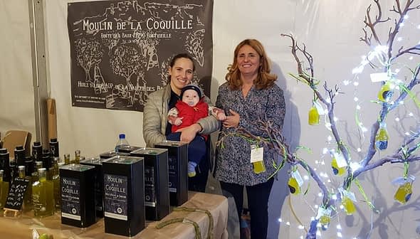 Two women and a baby at an olive oil stand with products displayed in a tent. - Olive Oil Times