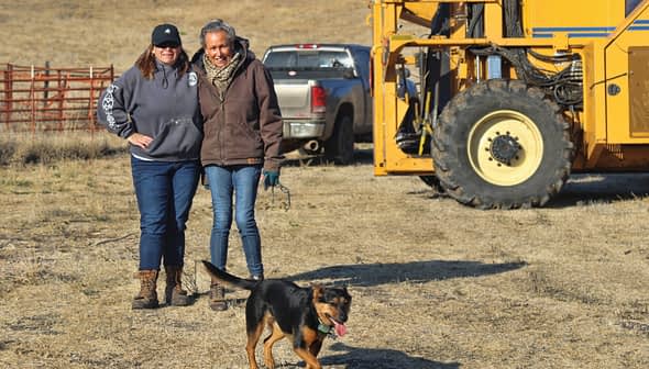 Two women standing next to a yellow machine with a dog running in front of them in an open field. - Olive Oil Times