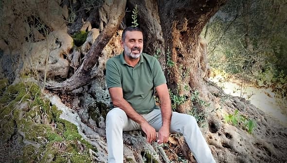 Man with a beard sitting on the roots of an olive tree, wearing a green shirt and light-colored pants. - Olive Oil Times