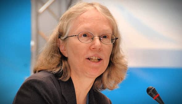 A woman with glasses speaking into a microphone at a conference setting. - Olive Oil Times